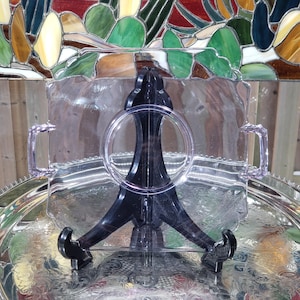 May include: A clear, rectangular serving tray with handles, resting on a black stand. The tray is made of clear plastic and sits on a silver tray with an ornate design. The background features a colorful stained-glass window.
