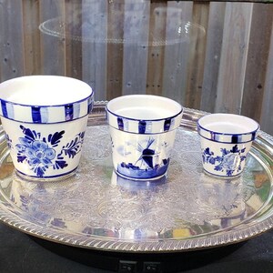May include: Three white ceramic flower pots with blue hand-painted floral and windmill designs, featuring blue striped rims. The pots are arranged in descending order of size and are placed on a large, round, silver-toned metal serving tray.