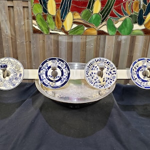 May include: A wall-mounted rack featuring four circular ceramic plates with blue and white floral patterns, each centered with a metal hook and a small ceramic knob. The plates are attached to a horizontal off-white wooden bar.