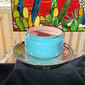 May include: A bright turquoise bowl with a smooth, light pink interior, resting on a round, polished silver-toned metal tray with an ornate, embossed rim. The bowl has a wide, cylindrical shape and a glossy finish, centred on the reflective tray.