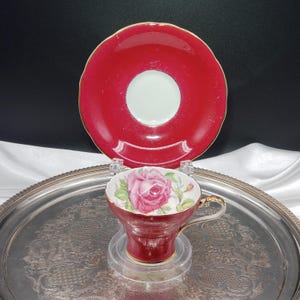 May include: A porcelain teacup and saucer set featuring a deep red glaze with gold trim. The teacup displays a detailed pink rose illustration on the interior and exterior, while the saucer has a solid red surface with a white centre circle.