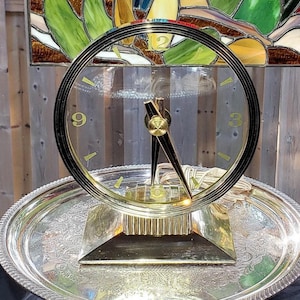 May include: A round analog clock with a clear glass face, gold-tone metal frame, and a square base. The clock features gold-tone hands and numerals at the 3, 9, and 12 positions, resting on a large, round, silver-tone metal serving tray.