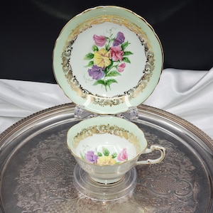 May include: A vintage teacup and saucer set. The set has a pale green border with gold filigree and a floral bouquet design. The teacup and saucer are displayed on a silver tray.