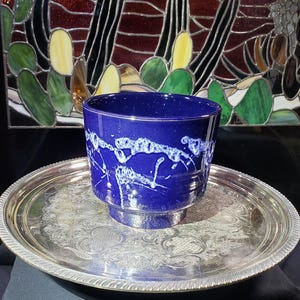 May include: A deep cobalt blue ceramic cup with a white, abstract, dripped glaze pattern around the centre. The cup sits centred on a round, polished silver-toned metal tray featuring an intricate, embossed floral and scrollwork design.