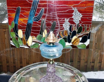 May include: An oil lamp with a clear glass chimney, a brass burner, a light blue glass font, and a purple patterned glass base. Behind the lamp is a rectangular stained glass panel depicting a desert landscape with cacti in red, blue, and green.