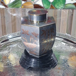 May include: A silver hexagonal tea caddy with a matching lid, decorated with etched floral and bamboo designs in copper and gold. The caddy sits on a black, tiered wooden base, displayed on a silver tray.
