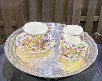 Pode incluir: Uma bandeja de servir circular em tom prateado com um padrão gravado ornamentado e uma borda estriada. Na bandeja, há uma xícara de chá de porcelana com pires correspondente, uma pequena leiteira e uma tigela, todas com estampa floral.