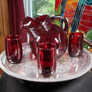 May include: A set of ruby red glassware consisting of a rounded pitcher with a handle and three matching tumblers. The glassware is arranged on a round silver-toned metal tray featuring an embossed floral and grape pattern along the rim.