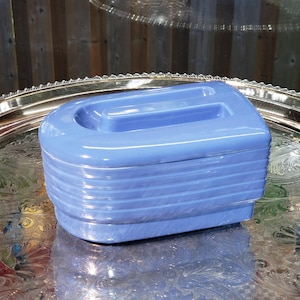 May include: A light blue, art deco style ceramic box with a handle on top. The box has horizontal ridges around the sides and sits on a silver tray. The box is a solid, opaque colour.