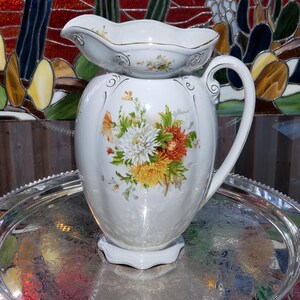 May include: A white porcelain jug featuring a scalloped rim with gold trim and a handle. The body is decorated with a vibrant floral arrangement of white, yellow, and orange chrysanthemums and green leaves, accented by delicate gold scrollwork.