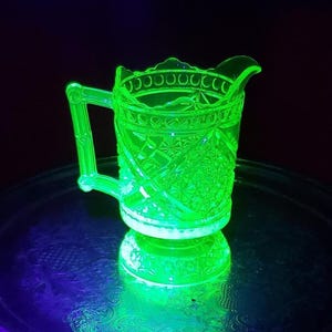 May include: A vibrant neon green glowing glass creamer pitcher featuring a detailed pressed pattern with geometric shapes and starburst motifs. The pitcher has a sturdy handle and a scalloped rim, illuminated under ultraviolet light to show its fluorescence.