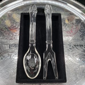 Peut inclure: Un ensemble composé d'une cuillère de service et d'une fourchette à deux dents en verre transparent. Les deux ustensiles présentent un manche à côtes verticales et sont ornés de motifs floraux gravés sur le cuilleron et les dents.