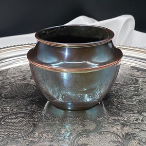 May include: A dark brown, metallic bowl with a smooth, rounded body and a distinct, flared rim. The bowl features a subtle, embossed floral pattern around its lower section and a polished copper-toned edge along the top rim, creating a contrast.
