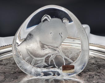 May include: A clear, triangular glass paperweight featuring a frosted, three-dimensional relief of a panda bear eating bamboo. The glass has smooth, rounded edges and a small, rectangular brown label attached to the lower right corner of the piece.