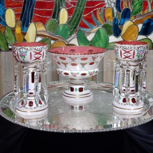 May include: A set of two white and red glass pedestal candle holders with hanging crystal prisms and a matching central glass bowl, all featuring hand-painted floral motifs and gold trim, arranged on a round silver-toned metal serving tray.