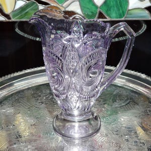 May include: A light purple glass pitcher featuring a pressed pattern of swirling scrolls, diamond shapes, and vertical ridges. The vessel has a scalloped rim, a sturdy handle on the right side, and a circular pedestal base, all crafted from translucent glass.