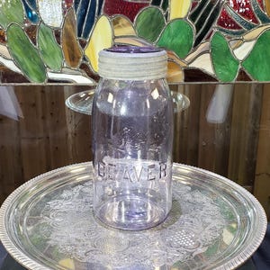May include: A clear glass jar with a light purple tint featuring the embossed word BEAVER on the front and a textured glass lid. The jar has a cylindrical body and a threaded neck, showcasing a distinct relief design of a beaver above the text.