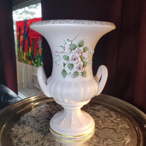 May include: A matte white ceramic urn-style vase featuring two side handles, a fluted base, and a wide rim with gold trim. The front is decorated with a raised, three-dimensional floral arrangement of pale pink roses, green leaves, and small blue flowers.