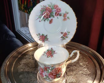 May include: A white porcelain teacup and matching saucer set featuring a delicate pattern of pink roses with green leaves. Both pieces have a scalloped edge and are finished with a thin gold trim along the rims and the teacup handle.