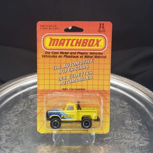 May include: A Matchbox M853 F150 Flareside toy truck in a yellow and blue color scheme, packaged on a yellow and orange card. The card reads "THE AUTOMOTIVE SUPERSTARS" and "FORD". The toy is in a clear plastic case.
