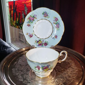 Peut inclure: Un ensemble tasse et soucoupe en porcelaine avec un fond bleu pâle, des bords festonnés et une bordure dorée. L'ensemble est orné de bouquets floraux colorés comprenant des fleurs roses, jaunes et violettes. La tasse possède une anse courbée.