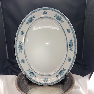 May include: An oval ceramic serving platter with a white base and a scalloped edge. The rim features a decorative border pattern in teal blue, consisting of repeating floral clusters and a delicate geometric line design around the centre.
