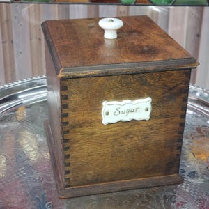 May include: A square wooden storage canister with a dark brown finish and visible finger joints at the corners. The lid features a white ceramic knob. A white ceramic label with a decorative border is attached to the front, displaying the word Sugar.