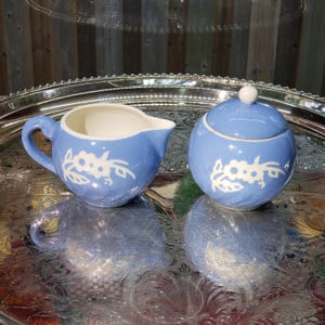 May include: A light blue creamer and sugar bowl set with white floral designs. The creamer has a handle and spout, while the sugar bowl has a lid with a small white knob. Both pieces are on a silver tray.