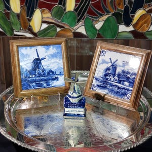 Peut inclure: Deux carreaux de céramique bleus et blancs encadrés avec des scènes de moulins à vent, et une petite figurine de moulin à vent. Les carreaux sont dans des cadres en bois. La figurine de moulin à vent est bleue et blanche. Les articles sont exposés sur un plateau argenté.