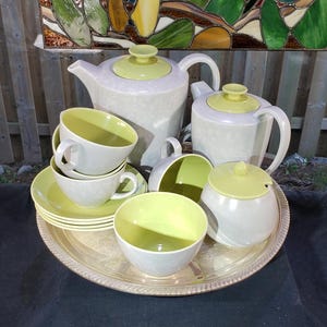 May include: A tea set featuring a large teapot, a smaller teapot, a sugar bowl with a lid, and several cups with saucers. The pieces are cream-colored with light green lids and interiors, all arranged on a round, metallic serving tray.