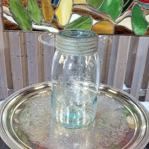 May include: A clear glass mason jar with a light blue tint and a threaded metal lid. The jar features embossed lettering on the front that reads Crown. The glass has a smooth, cylindrical body with a slightly tapered neck and a wide mouth opening.