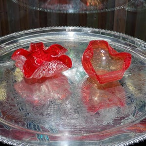 Peut inclure: Deux bols en verre aux bords ondulés sont posés sur un plateau en argent. Le bol de gauche est rouge profond avec un motif blanc tacheté. Le bol de droite est rouge orangé translucide avec une texture à bulles sur toute la surface.