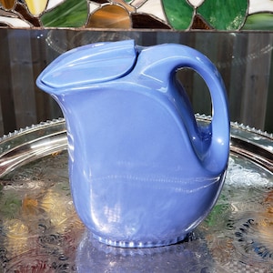May include: A periwinkle blue ceramic pitcher with a smooth, glossy finish, featuring a curved handle, a wide spout, and a matching lid. The pitcher has a rounded, bulbous body with a subtle, flared base, showcasing a solid, uniform colour throughout.
