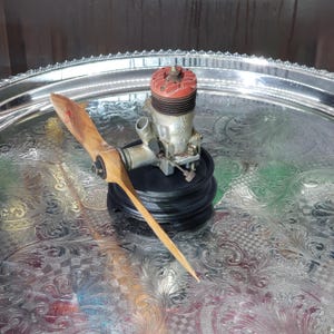 May include: A vintage McCoy model aeroplane engine with a red cylinder head and a silver metal body, attached to a wooden propeller. The engine is mounted on a black circular base, featuring visible mechanical components and metal hardware.