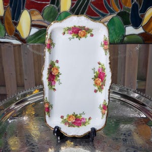 May include: A rectangular white porcelain serving tray with scalloped edges and a gold-trimmed rim. The tray features a floral pattern of red, yellow, and pink roses with green leaves arranged in clusters along the centre of each side.