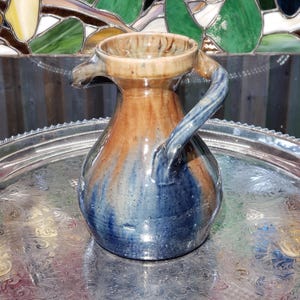 May include: A ceramic pitcher featuring a rounded body and a single handle. The glaze displays a blend of deep blue at the base transitioning into warm tan and brown tones toward the rim. The surface has a glossy finish with visible color drips.