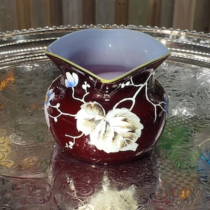 May include: A small, deep burgundy ceramic creamer with a light lavender interior and a gold-toned rim. The exterior features hand-painted white and gold leaf motifs with small blue floral accents, showcasing a rounded body and a wide pouring spout.