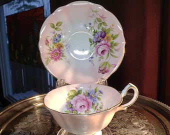 May include: A pale pink porcelain teacup and matching saucer set featuring a scalloped edge with gold trim. The set is decorated with a colourful floral pattern including pink roses, blue flowers, and green leaves on both the cup and saucer.