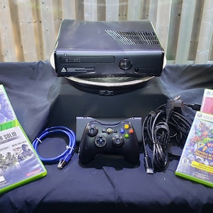 May include: A black Xbox 360 console with a black controller, a blue ethernet cable, and a black power cable. There are five video game cases, including "American Idol", "Metal Gear Solid", "Viva Piñata", and two other games.