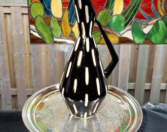 May include: A black ceramic pitcher with a glossy finish and vertical white dash patterns, featuring a tapered neck and a curved handle, resting on a round silver-toned metal tray with an intricate embossed floral pattern along the rim.