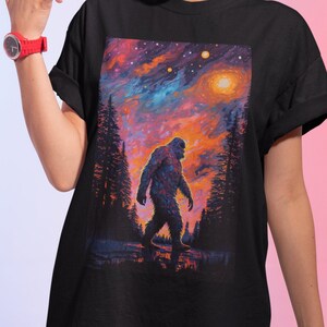 May include: Black t-shirt with a colorful graphic print of a Bigfoot walking through a forest under a starry sky.