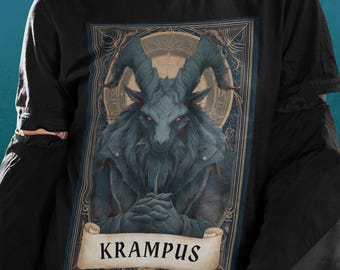Puede incluir: Camiseta negra con una ilustración detallada de Krampus, una figura con cuernos y expresión severa, enmarcada en un marco ornamentado. El diseño incluye una pancarta con el nombre "KRAMPUS". La camiseta tiene mangas largas.