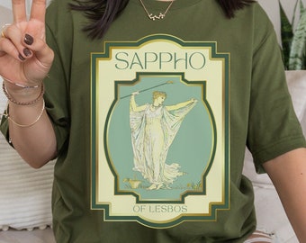 May include: Olive green t-shirt featuring a framed illustration of Sappho of Lesbos. The artwork depicts a woman in a flowing gown holding a staff. The text "SAPPHO OF LESBOS" is printed above and below the image in a gold-colored font.