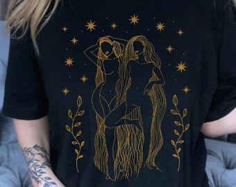 Puede incluir: Una camiseta negra con una ilustración de contorno dorado de dos mujeres con cabello largo y fluido, rodeadas de estrellas y enredaderas florales.