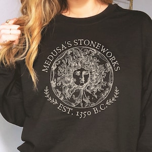 Peut inclure: Sweat-shirt noir avec un imprim&eacute; graphique blanc de Medusa's Stoneworks, repr&eacute;sentant une image stylis&eacute;e de la t&ecirc;te de M&eacute;duse avec des serpents pour cheveux, entour&eacute;e d'une couronne de laurier. Le texte indique "Medusa's Stoneworks Est. 1350 B.C."