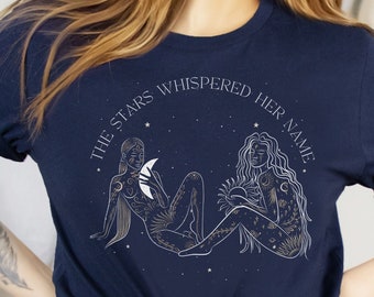 May include: Navy blue t-shirt featuring a celestial design with two figures, one holding a crescent moon and the other the sun. The text "THE STARS WHISPERED HER NAME" is arched above the figures. The shirt has rolled-up sleeves.