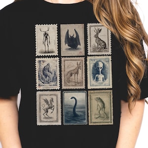 Può includere: T-shirt nera con nove francobolli in stile vintage raffiguranti criptidi e creature mitologiche. I francobolli presentano illustrazioni di un Bigfoot, un pipistrello, un jackalope, un serpente marino e altre creature misteriose.