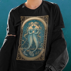 Puede incluir: Camiseta negra con una ilustraci&oacute;n vintage de dos sirenas con colas azules y detalles dorados. Las sirenas se miran y sus colas est&aacute;n entrelazadas. La ilustraci&oacute;n est&aacute; rodeada de un marco dorado con detalles ornamentados.