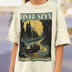 Peut inclure: T-shirt crème avec une illustration de style vintage d'une scène de rivière, avec le texte "RIVER STYX" et "rafting adventures". Le graphisme représente un bateau avec des passagers, des falaises sombres et un ciel jaune.