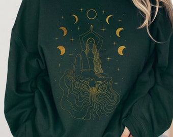 Puede incluir: Una sudadera verde oscuro con un gráfico dorado de una mujer con el pelo largo, rodeada de lunas y estrellas. El gráfico es una ilustración estilizada de una mujer en una postura de yoga.